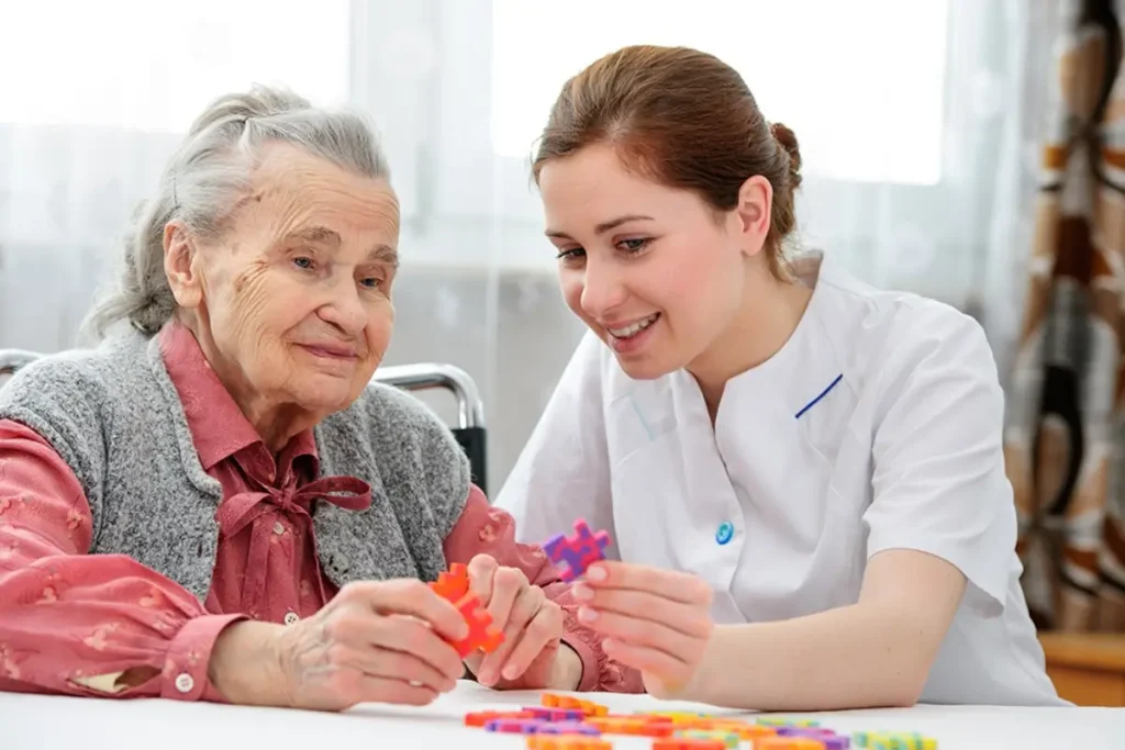 An elderly woman smiles at a caregiver who gently rests a hand on her shoulder, both sharing a moment of connection.