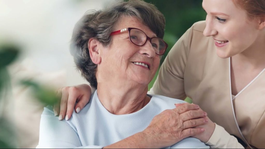 An elderly woman smiles at a caregiver who gently rests a hand on her shoulder, both sharing a moment of connection