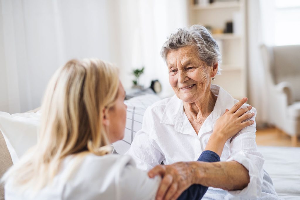 An elderly woman smiles warmly while being comforted by a caregiver, both sitting together indoors.