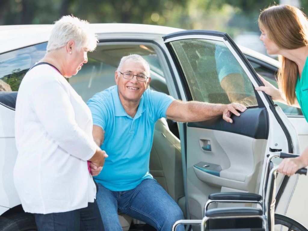 An elderly man is assisted by two women as he gets out of a car, with a wheelchair nearby.