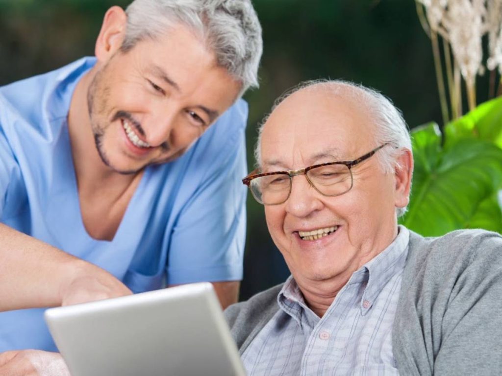 Smiling caregiver in blue scrubs with gray-haired elderly man in glasses looking at tablet together outdoors.