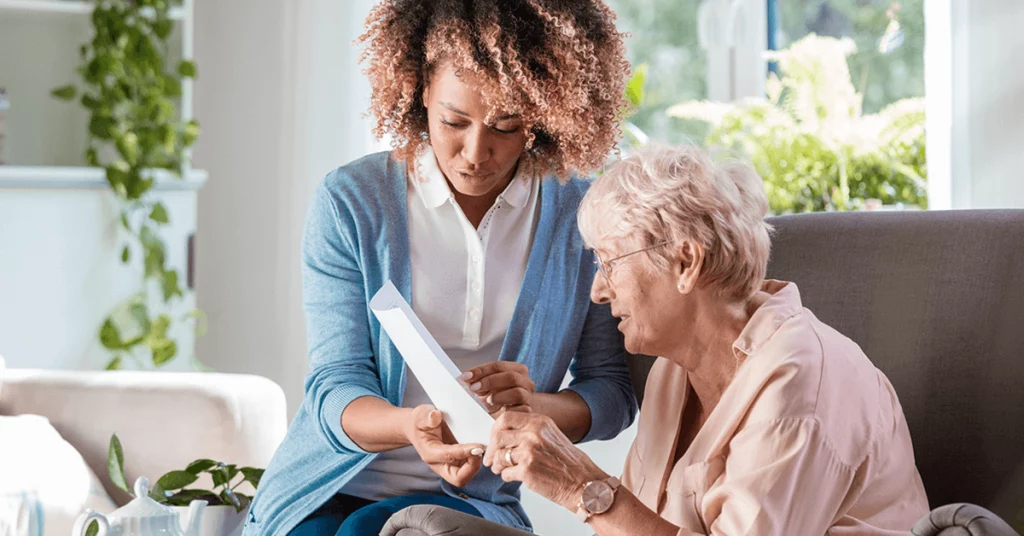 A caregiver showing a document to an elderly woman, both engaged in a thoughtful conversation in a bright, cozy setting.