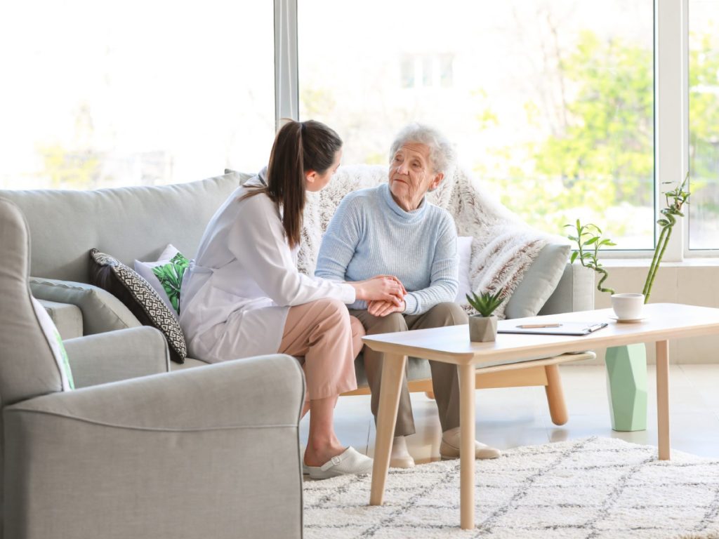 Young woman in white sitting with elderly woman in blue sweater on couch by large window in bright living room.
