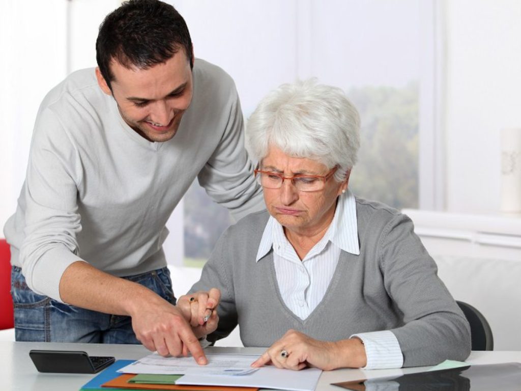 Young man helping elderly woman with glasses review documents at desk with laptop and papers in bright room.
