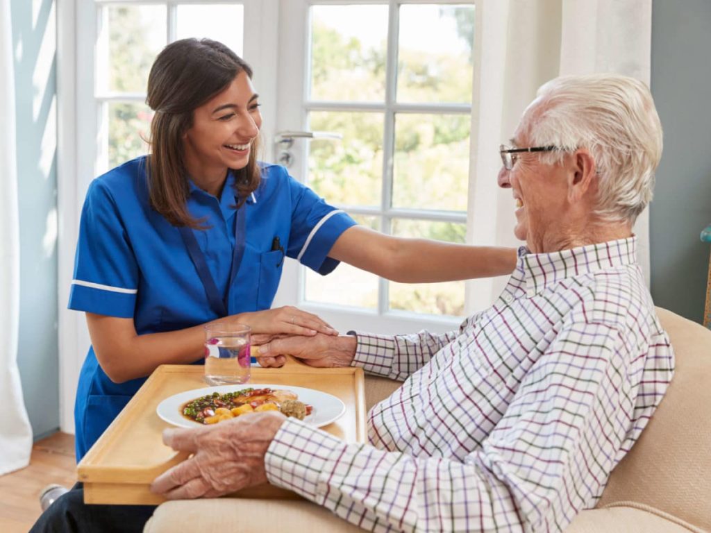 Young female caregiver in blue uniform helping elderly man with glasses eat meal at table by bright window
