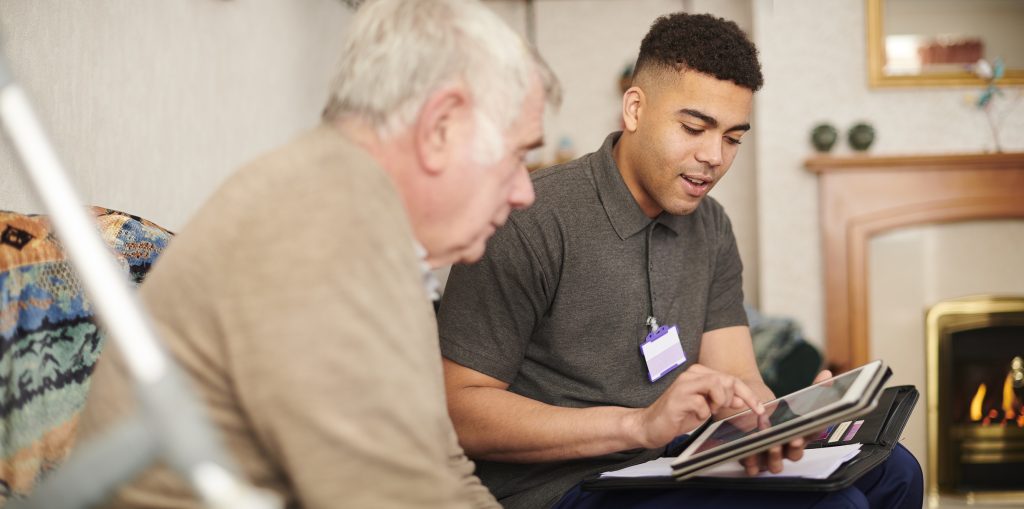 A caregiver helping an elderly man with a tablet, while sitting together in a cozy living room.