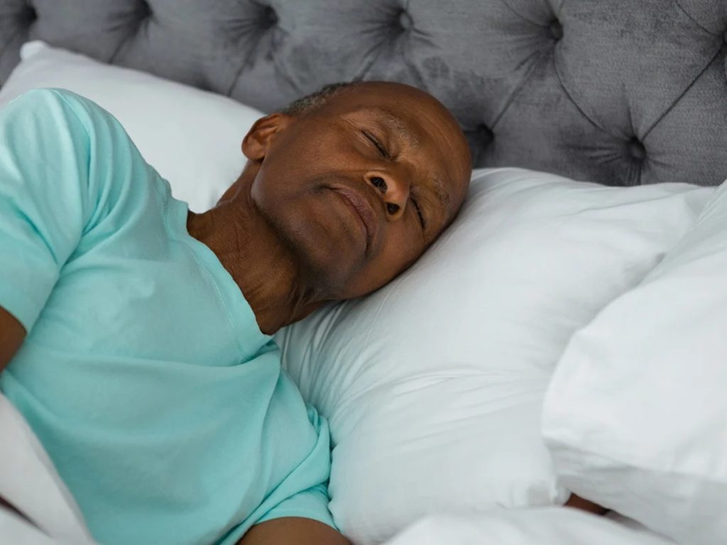 Elderly Black man in light blue shirt lying peacefully in bed with white pillows and gray headboard smiling