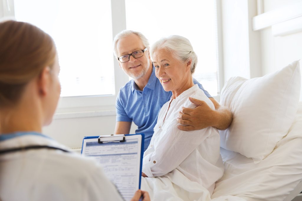 An elderly couple smiling as they speak with a healthcare professional, sitting together in a bright room.
