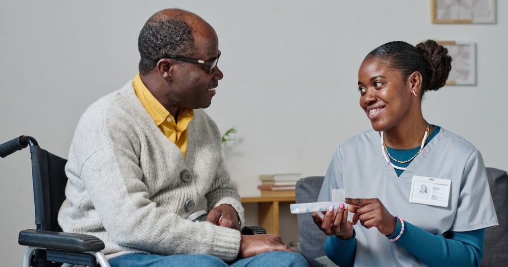 A caregiver handing medication to an elderly man in a wheelchair, both smiling and engaging in conversation.