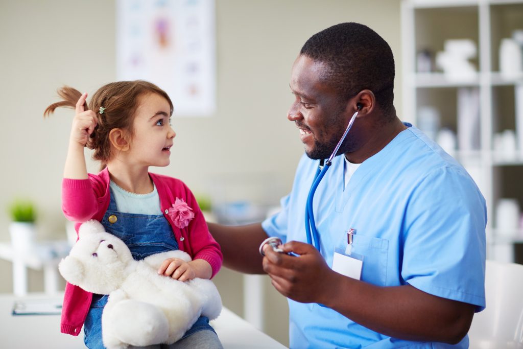 A young girl playfully talking with a doctor while holding a teddy bear, both smiling in a pediatric clinic.
