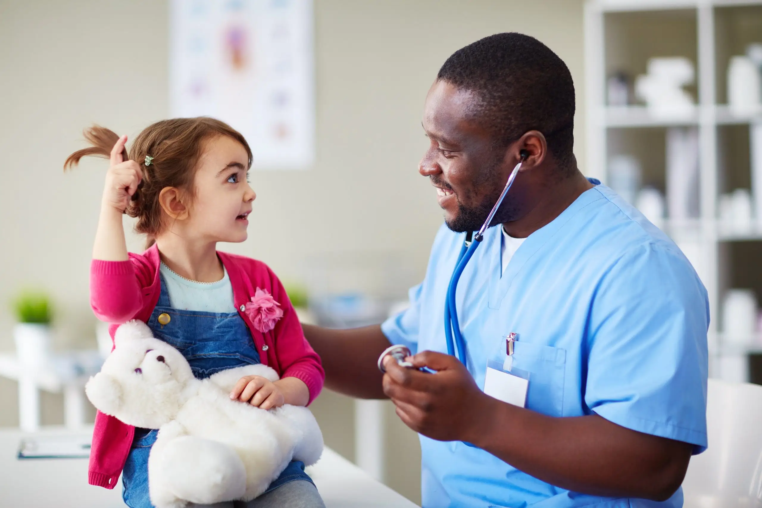 A young girl playfully talking with a doctor while holding a teddy bear, both smiling in a pediatric clinic.