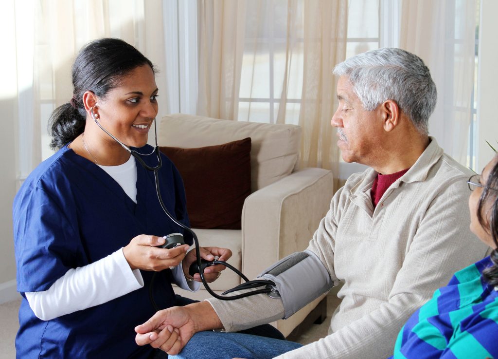 A nurse taking the blood pressure of an elderly man, while another person sits beside them, both in a well-lit room.