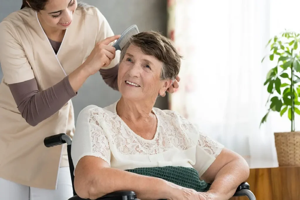 A caregiver combing the hair of an elderly woman in a wheelchair, both smiling in a bright, cozy room.
