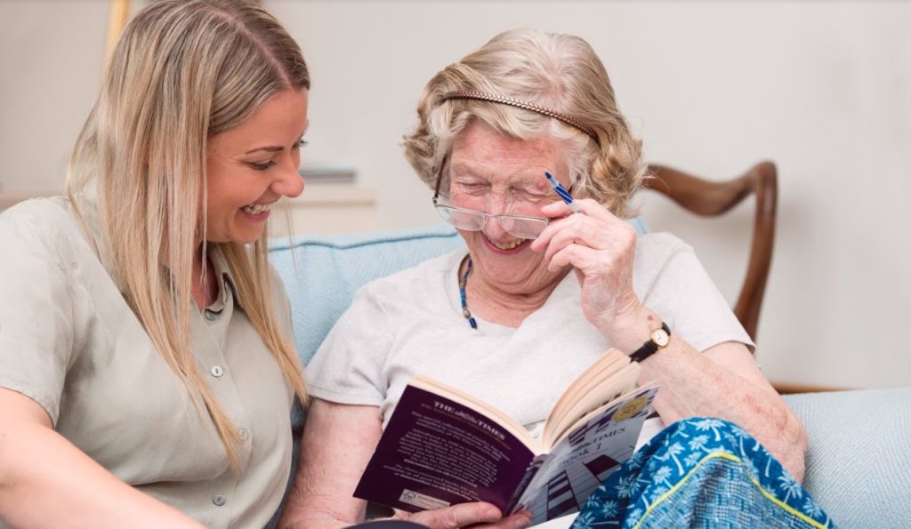 A caregiver and elderly woman smiling while reading a book together, with the woman adjusting her glasses.