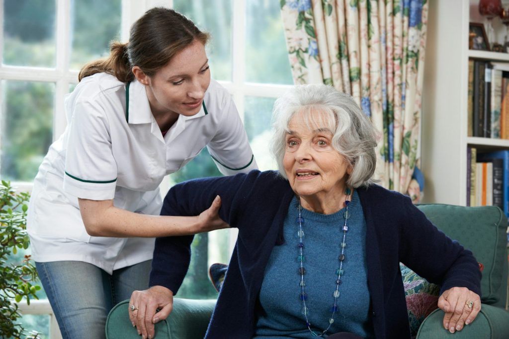 A caregiver assisting an elderly woman to stand up from a chair, both smiling and engaging in conversation.