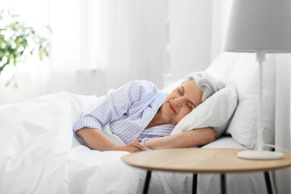 Elderly woman with gray hair sleeping peacefully in bed wearing blue striped pajamas in bright bedroom setting