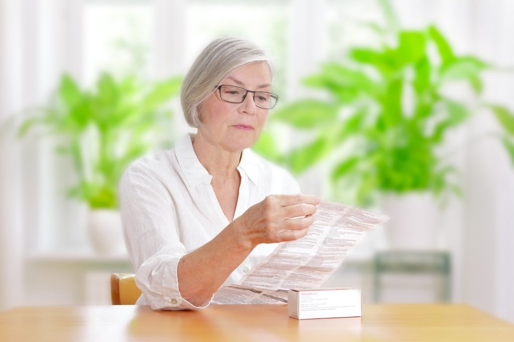An elderly woman reading the instructions on a medication package, sitting at a table in a well-lit room.