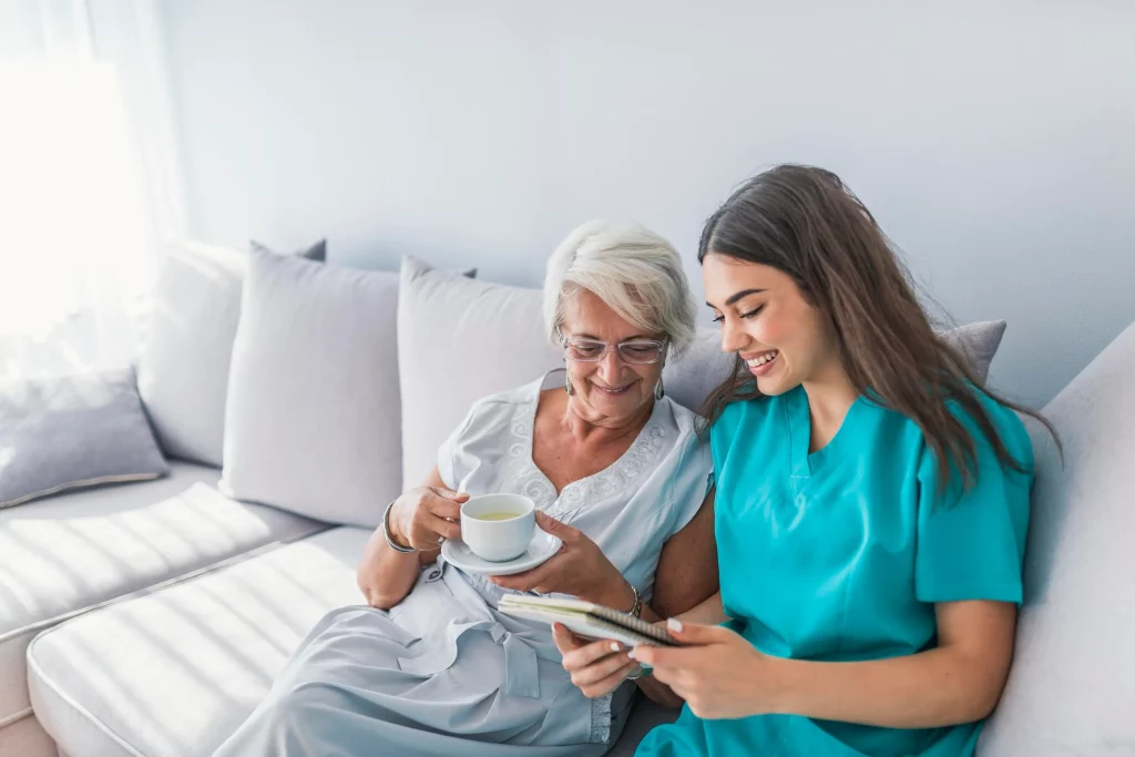 Caregiver and elderly woman smiling together on a couch, sharing tea and looking at a notebook.
