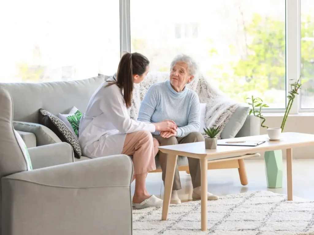 A caregiver sitting with an elderly woman on a couch, both smiling and holding hands in a bright living room.