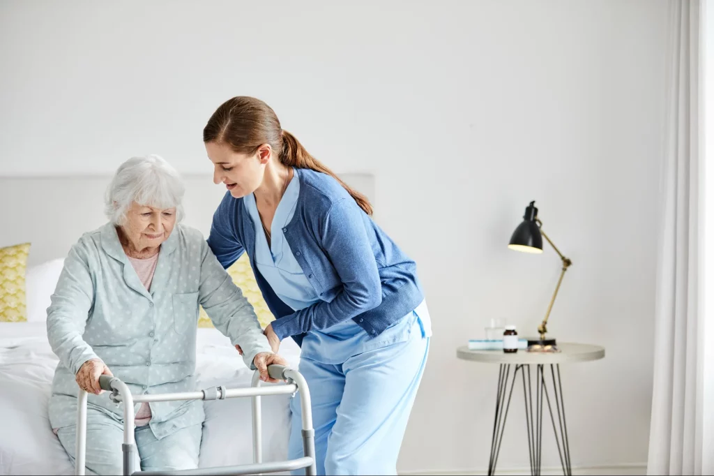 Young female caregiver in blue scrubs helping elderly woman with walker stand up in bright modern room.