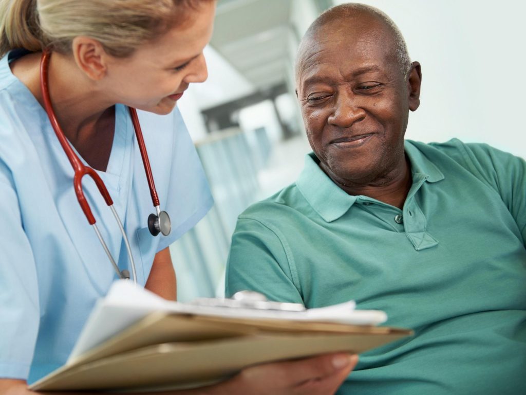 Female doctor with stethoscope reviewing medical chart with smiling elderly Black male patient in green shirt.