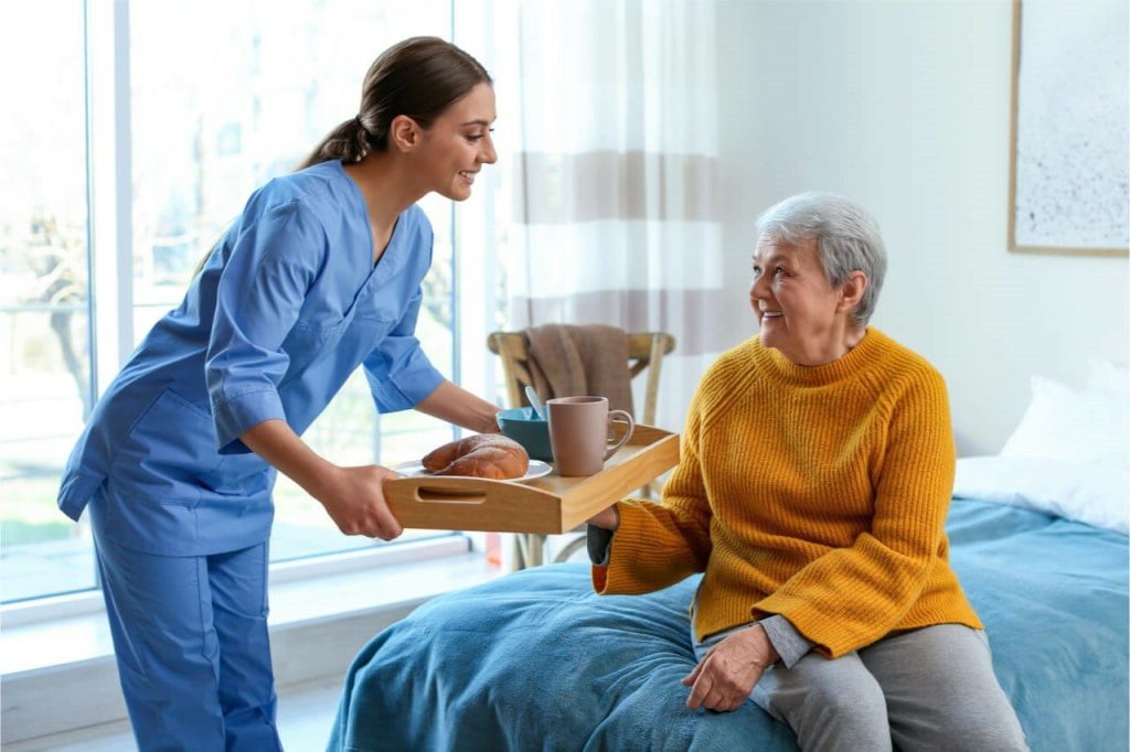 A caregiver in blue scrubs serving breakfast to an elderly woman in a yellow sweater in a cozy bedroom.